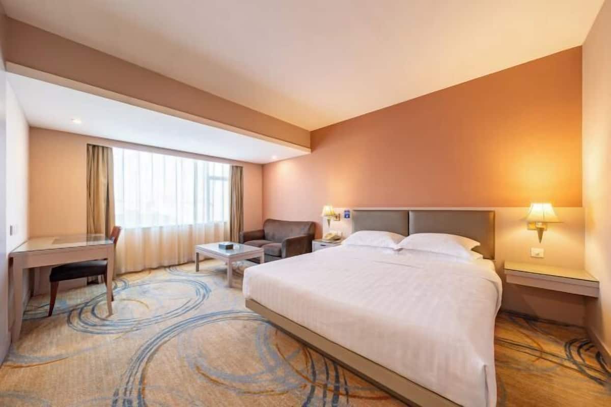 A hotel room with a large bed, a desk, a chair, a small table, and a nightstand with a lamp.