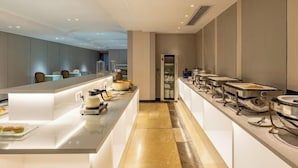 A modern hotel kitchen with a long counter, buffet setup, and seating area. - Dron Hotel (Dongguan)