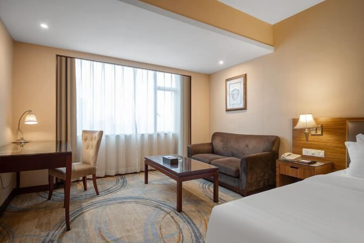 Ein Hotelzimmer mit einem Bett, einem Schreibtisch, einem Sessel, einer Couch, einem Nachttisch und einem großen Fenster mit Vorhängen.