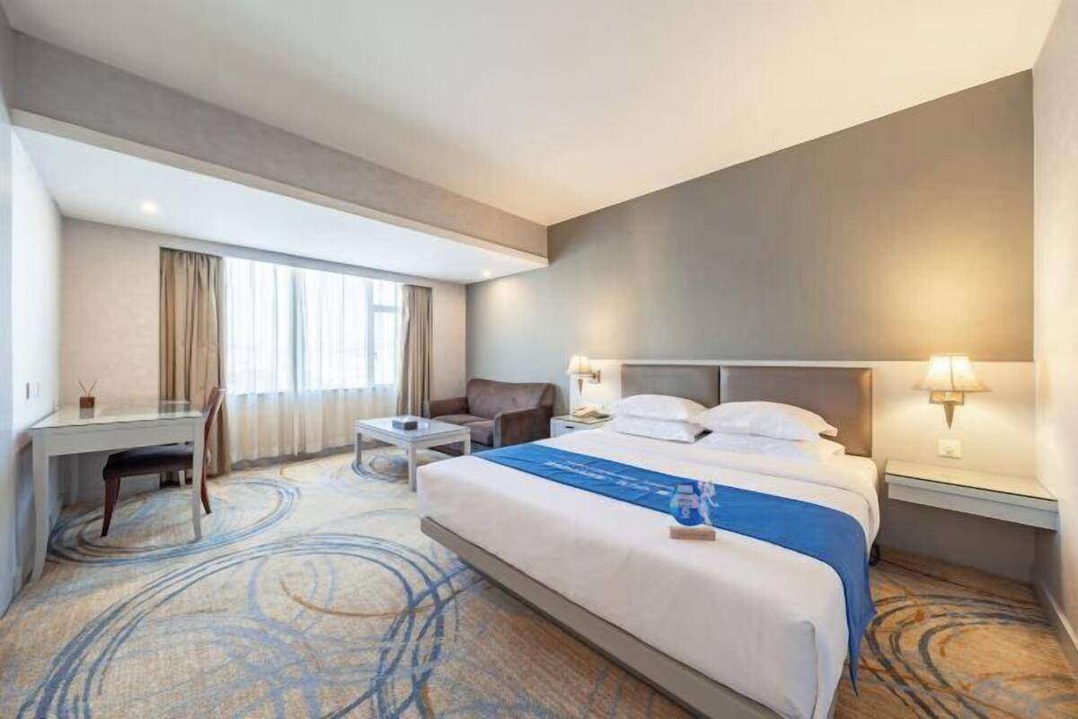 A hotel room with a large bed, a desk, a chair, a small table, and a nightstand with lamps.