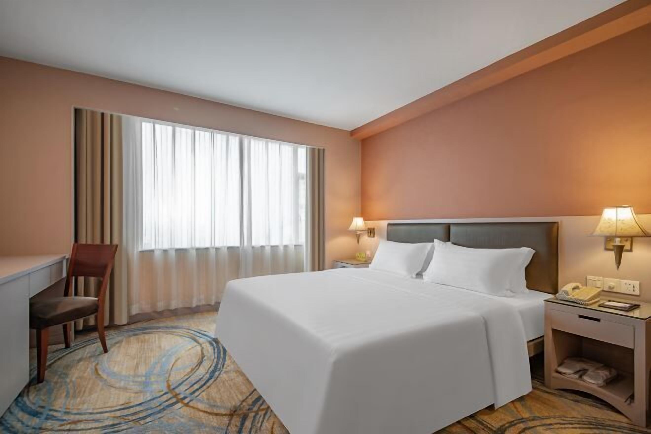 A hotel room with a large bed, a desk, a chair, a nightstand with a lamp, and a window with curtains.