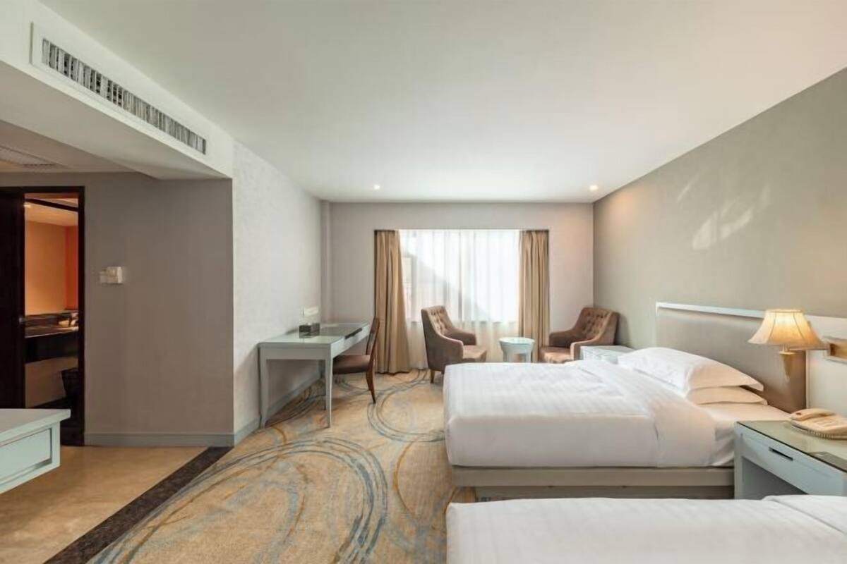A hotel room with two beds, a desk, a chair, a small table, and a lamp.