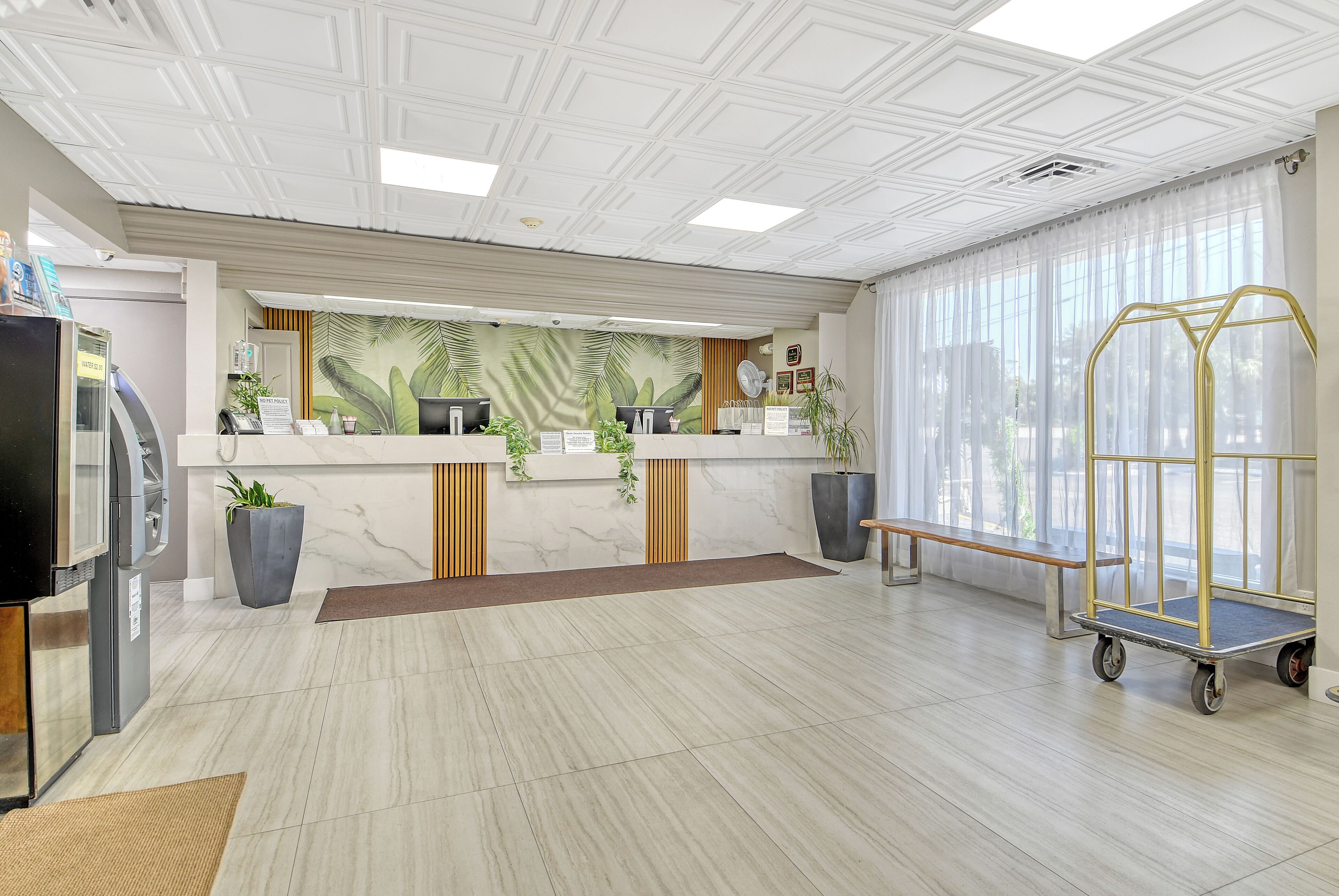 A reception area with a marble counter, two computer stations, a large window with sheer curtains, a bench, and a cart.