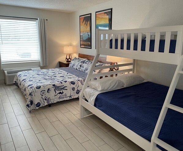 A room with two bunk beds, a bed with a blue quilt, a window with blinds, a wall-mounted lamp, and a framed picture of Sarasota, Florida. - Heated Pool, King Bed, Bunk Bed, Huge TV, Marina, Tiki Bar & Grill (Sarasota)