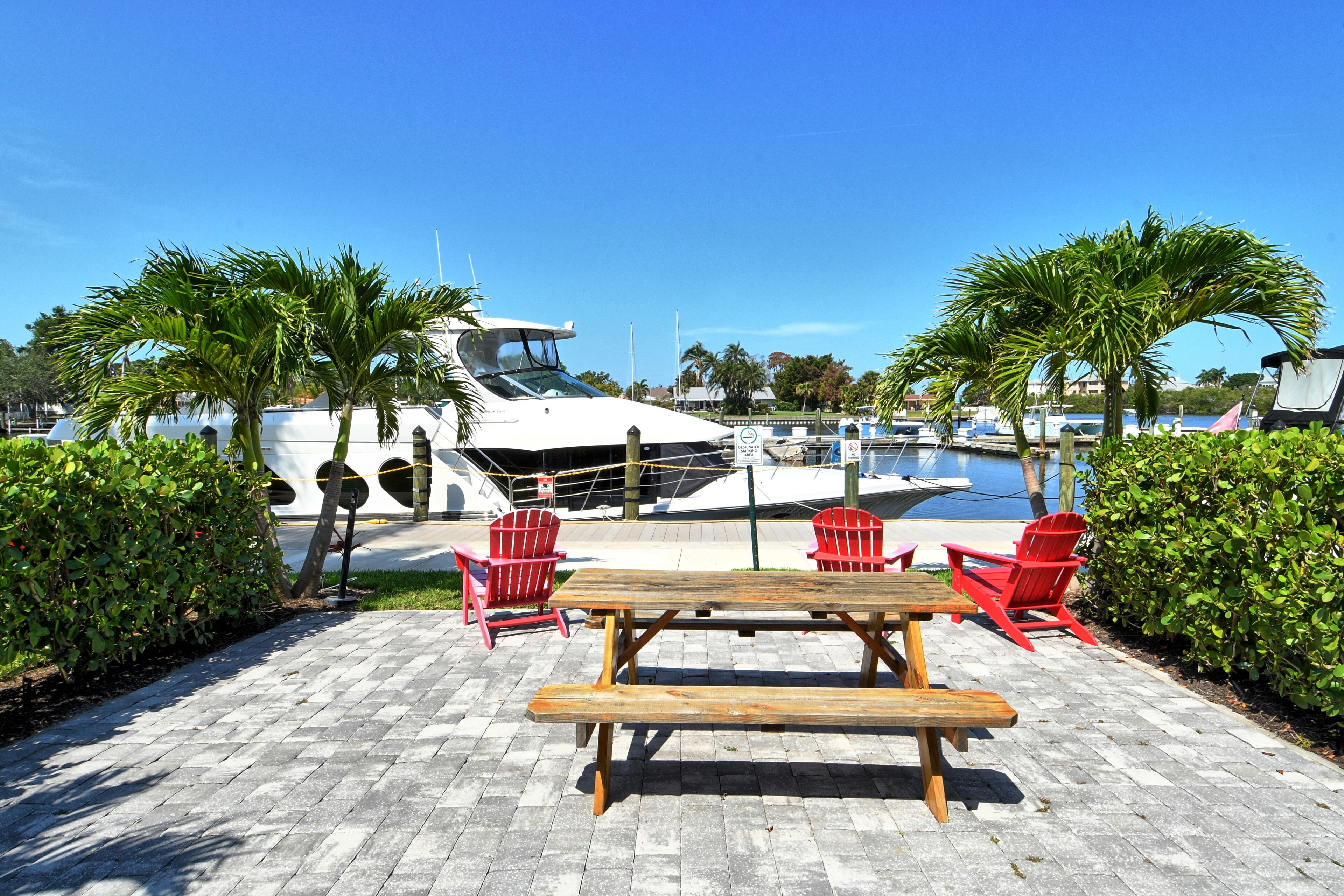 A wooden picnic table with benches, surrounded by red chairs, set on a paved area with palm trees and a marina in the background.