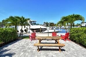 A wooden picnic table with benches, surrounded by red chairs, set on a paved area with palm trees and a marina in the background. - Heated Pool, King Bed, Bunk Bed, Huge TV, Marina, Tiki Bar & Grill (Sarasota)