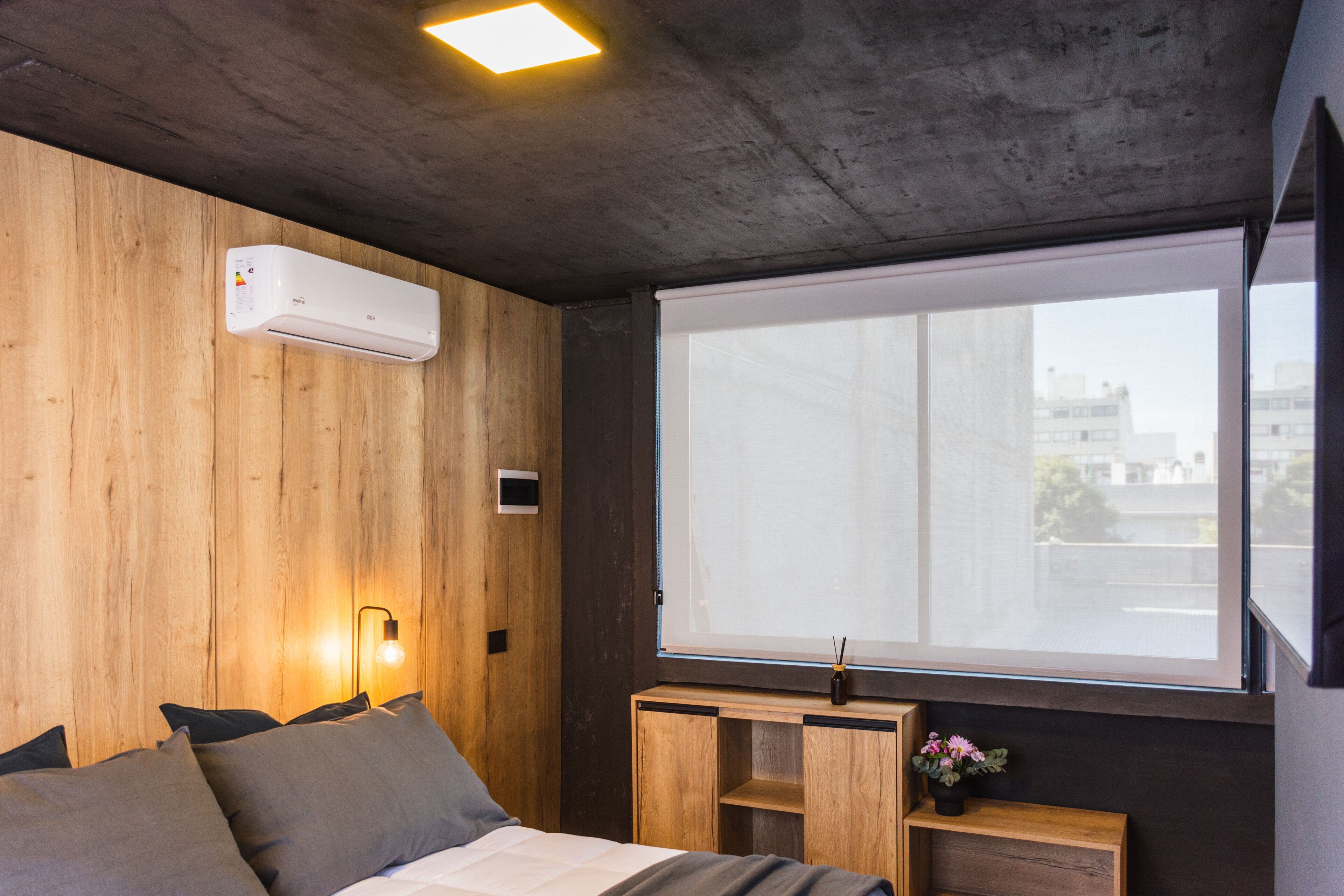 Una camera da letto moderna con una parete in legno, un'ampia finestra, un letto con cuscini, un comodino con un vaso e un condizionatore d'aria.