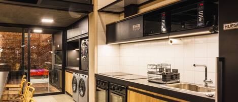 Una cocina moderna con electrodomésticos de acero inoxidable, horno empotrado y pileta. En la zona contigua hay sillas de mimbre y una mesa de comedor.