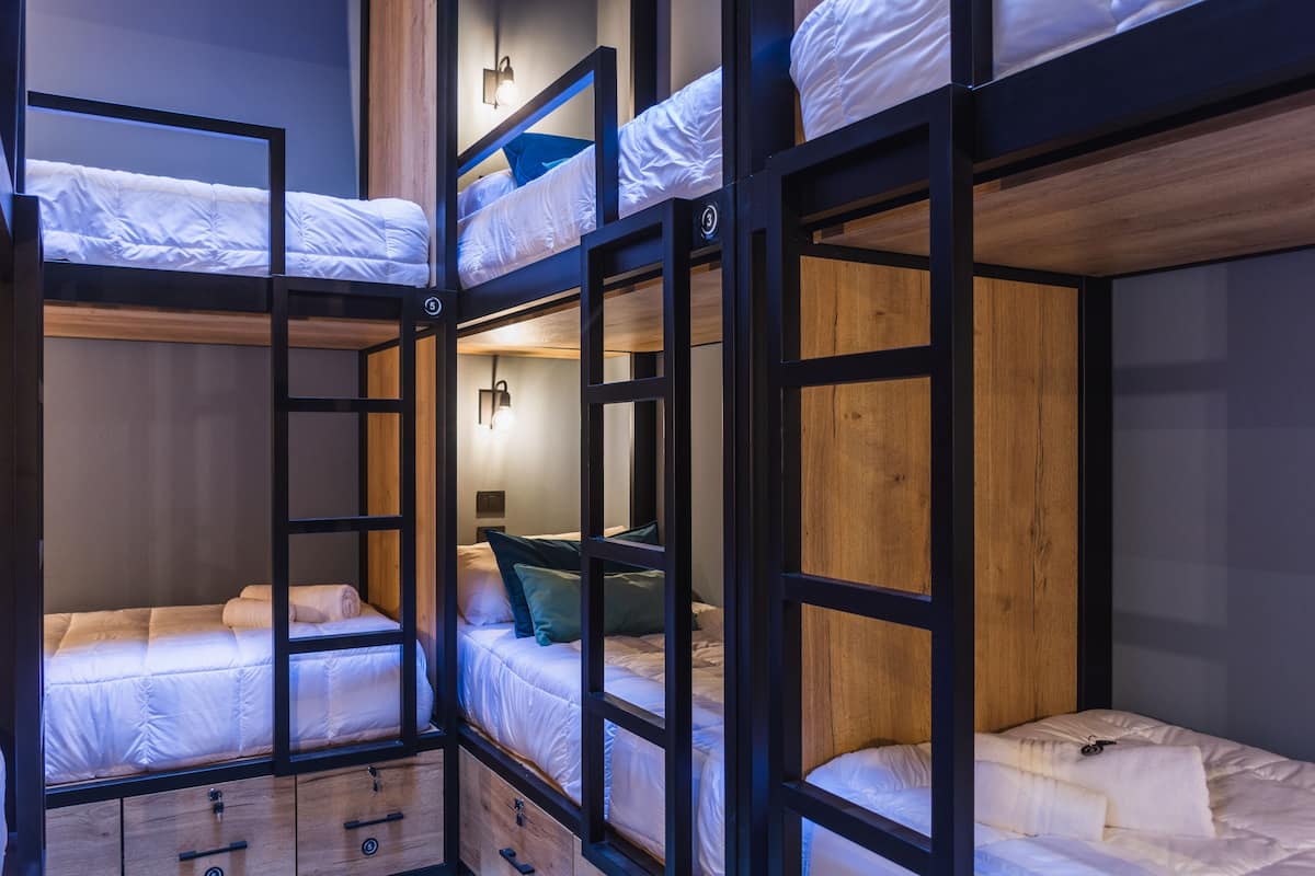 A bunk bed room with wooden frames, white bedding, and a small desk with drawers.