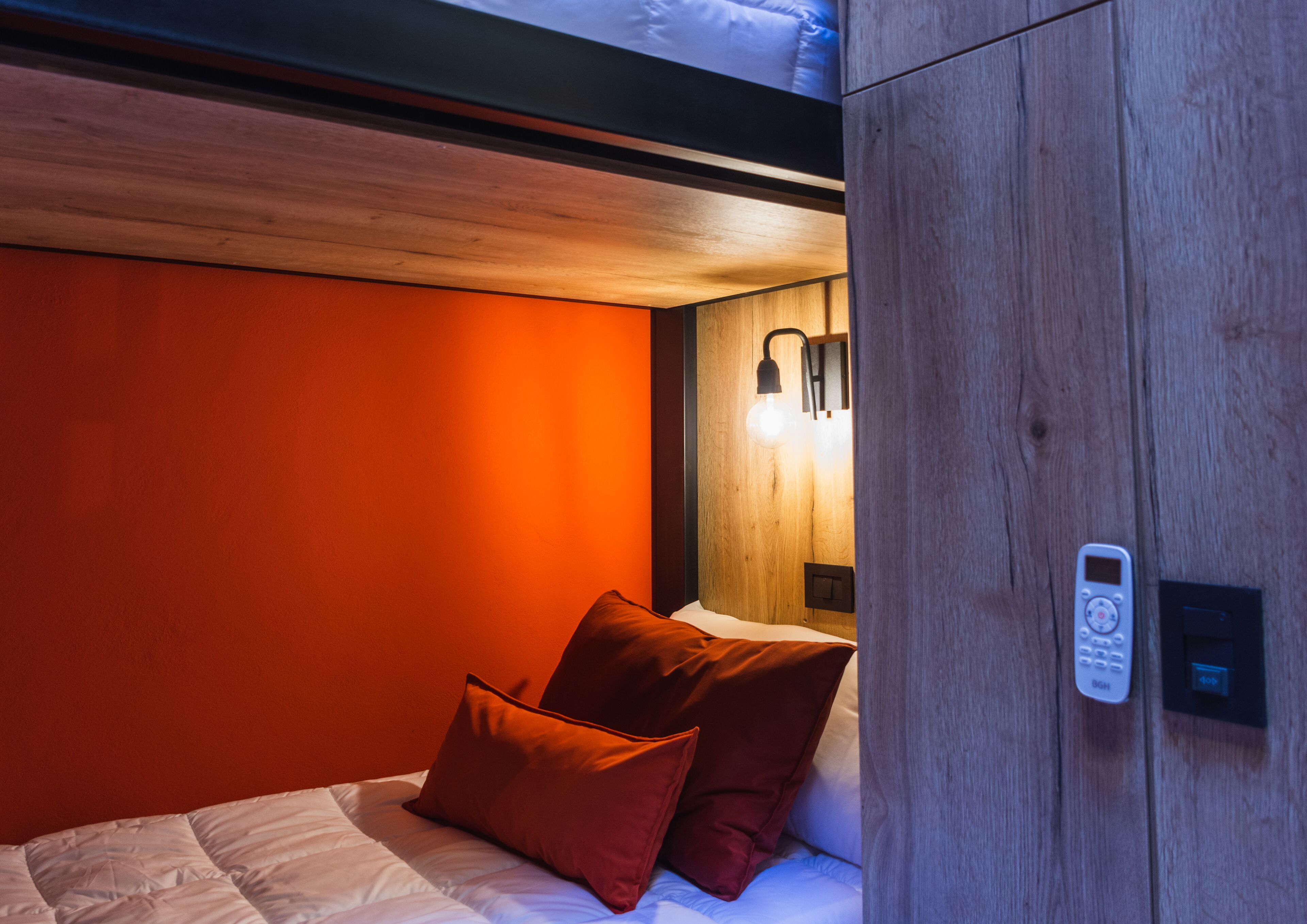 Un dormitorio con cabecera de madera, una cama con ropa de cama blanca y dos almohadas rojas. Hay una lámpara de pared y un panel de control en la pared.