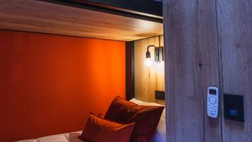 Un dormitorio con cabecera de madera, una cama con ropa de cama blanca y dos almohadas rojas. Hay una lámpara de pared y un panel de control en la pared.