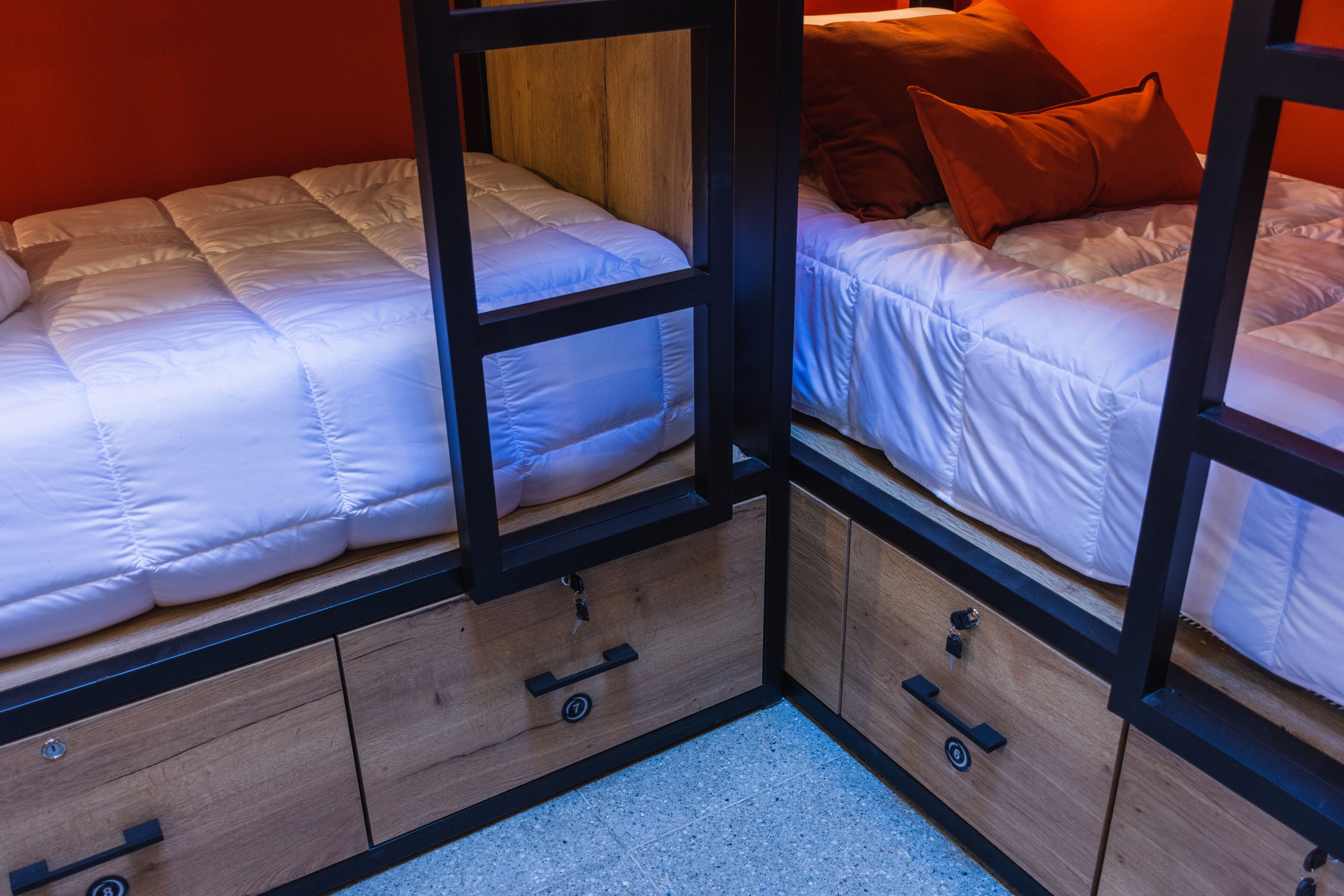 A room with two beds, each with a wooden frame and a white mattress. There are wooden drawers under the beds and a small rug on the floor.