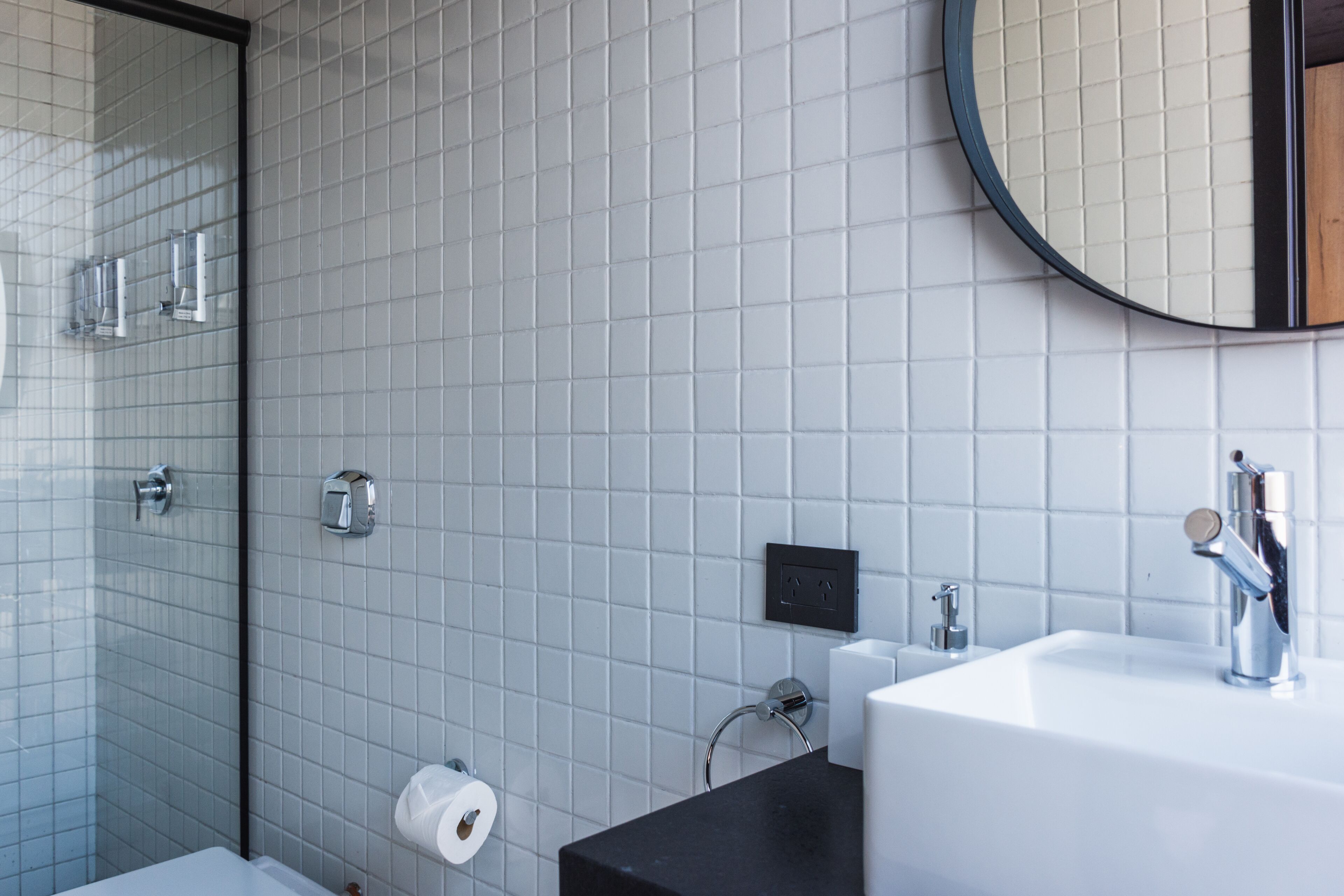 Un baño moderno con pared de azulejos blancos, encimera negra, lavamanos blanco, espejo redondo y mampara de ducha de vidrio.