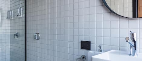 Un baño moderno con pared de azulejos blancos, encimera negra, lavamanos blanco, espejo redondo y mampara de ducha de vidrio.