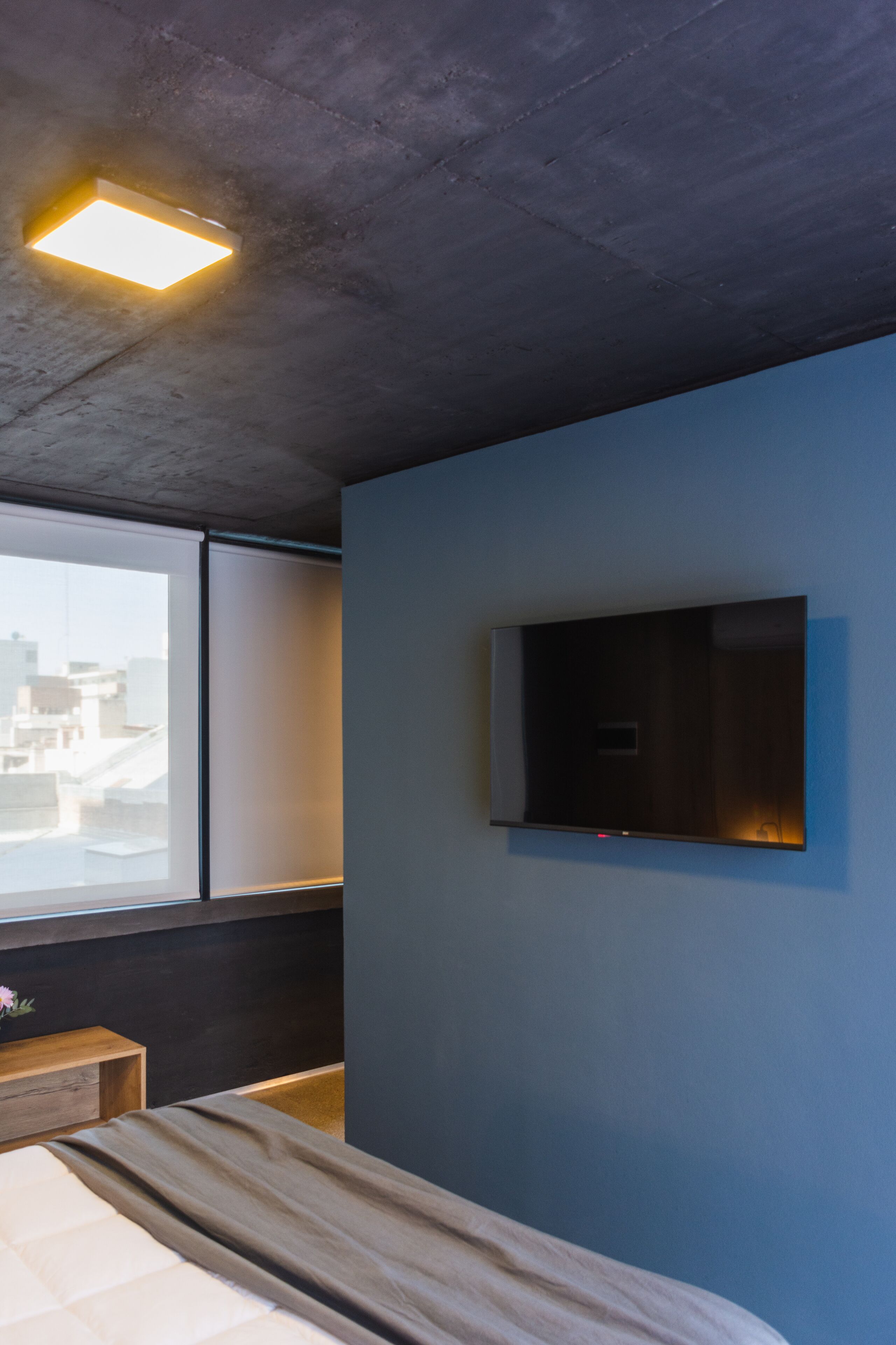 Habitació d'hotel moderna amb un llit, televisió de pantalla plana penjada a la paret, finestra amb vista a edificis i un llum al sostre.
