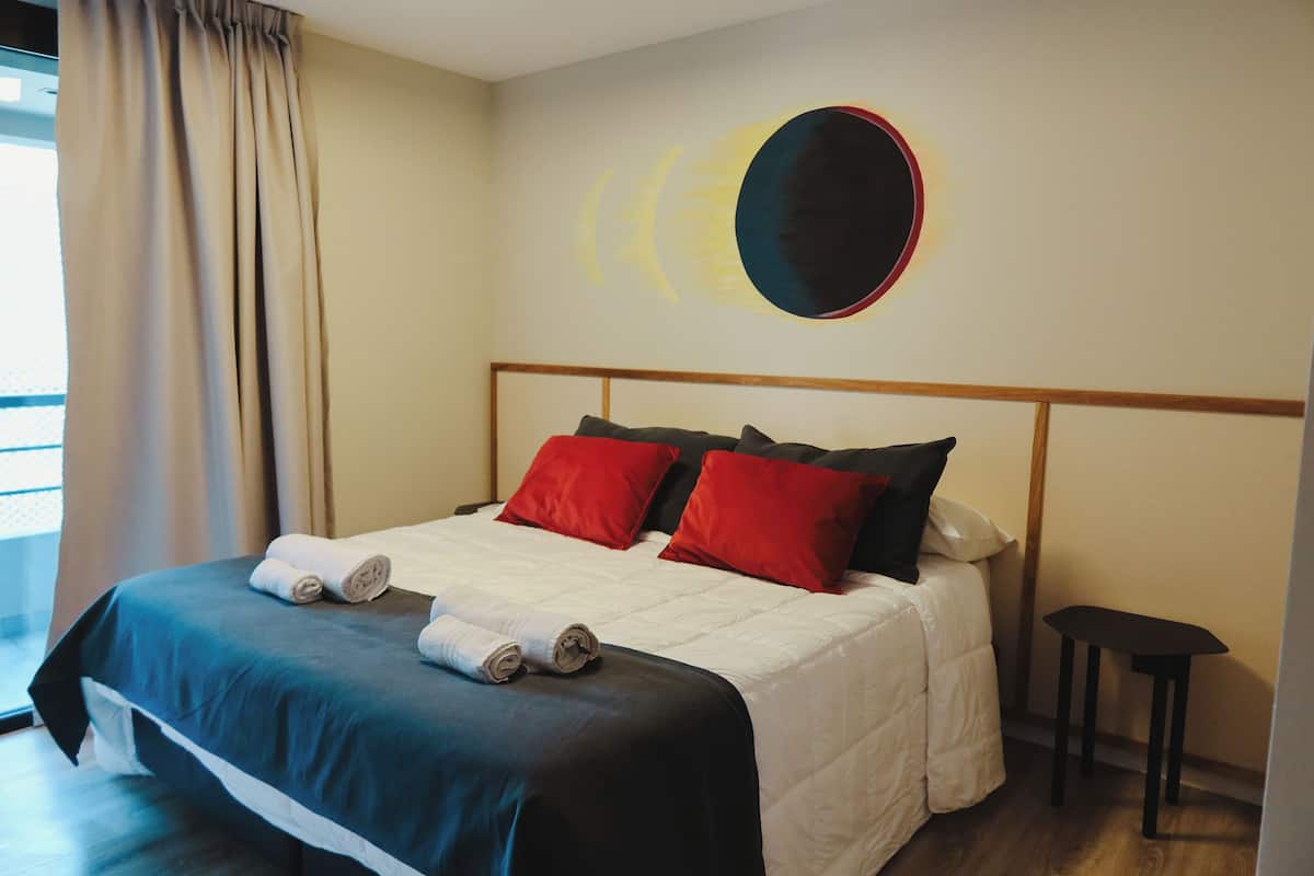 A hotel room with a bed, a headboard, a wall art piece, a window with curtains, and a small table.