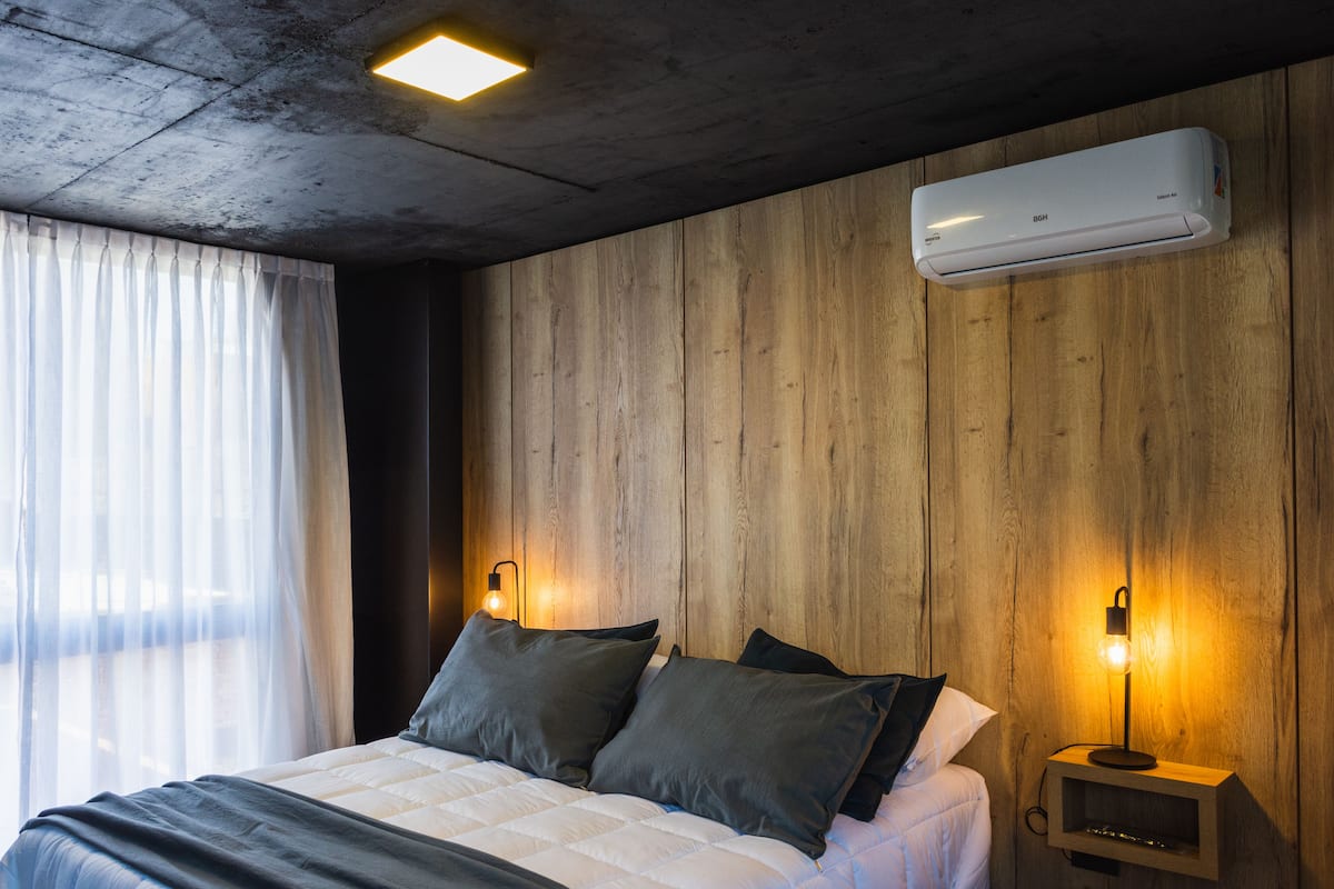 A bedroom with a wooden wall, a bed with pillows, a bedside table with a lamp, and an air conditioning unit.