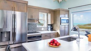 A modern kitchen with stainless steel appliances, a large island with a fruit bowl, and a view of the ocean through a window. - Chalk Sound Waterfront Cottage: Pool & Kayak (Providenciales)