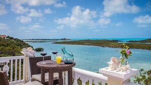 A balcony with wicker furniture, a table, and a view of a turquoise sea and green landscape. - Chalk Sound Waterfront Cottage: Pool & Kayak (Providenciales)