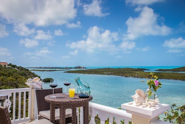 A balcony with wicker furniture, a table, and a view of a turquoise sea and green landscape.