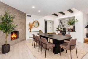 A modern dining area with a central table, chairs, a fireplace, and a kitchen in the background.