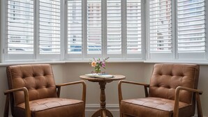 Dos sillones de cuero marrón con armazón de madera, una mesita redonda de madera con un jarrón de flores y una ventana con persianas blancas.