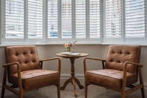 Two brown leather chairs with wooden frames, a small round wooden table with a vase of flowers, and a window with white blinds.