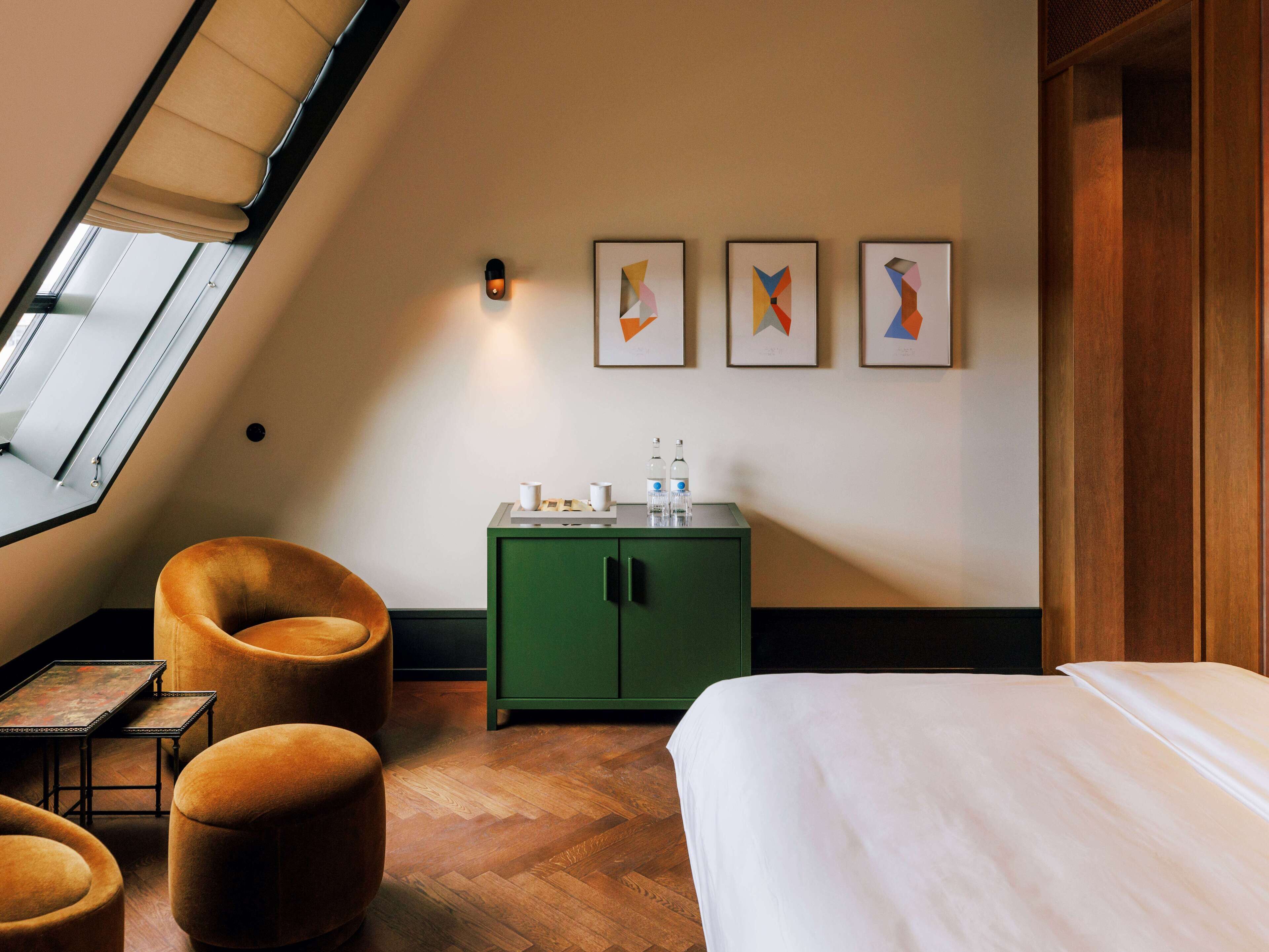 A hotel room with a sloped ceiling, a green minibar, a bed, a brown armchair, and a small table.