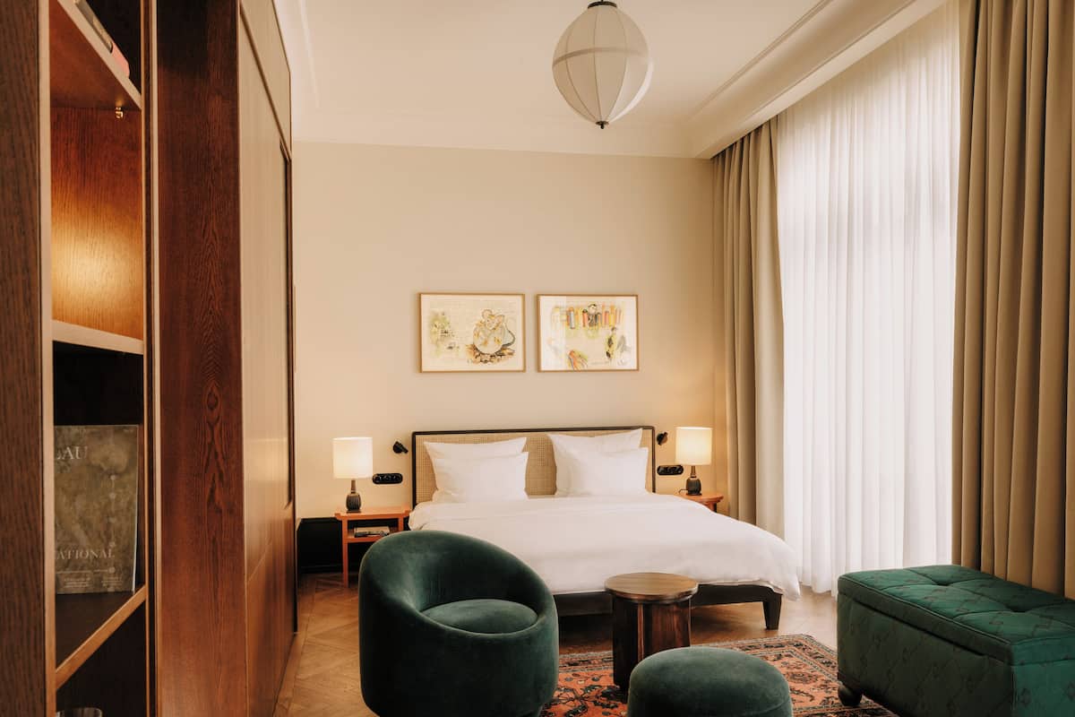 Une chambre d’hôtel avec un grand lit, deux tables de chevet avec des lampes, un fauteuil vert, un pouf vert, une armoire en bois et deux tableaux encadrés au mur.