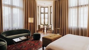 A hotel room with a bed, a green armchair, a small table, a lamp, and a view of the city through large windows. - Château Royal Berlin (Berlin)