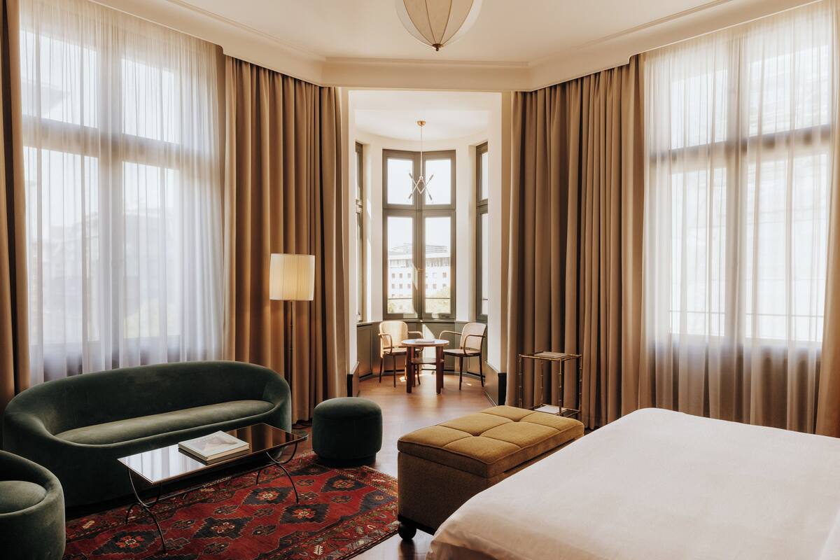 Une chambre d’hôtel avec un lit, un fauteuil vert, une petite table, une lampe et une vue sur la ville grâce à de grandes fenêtres.