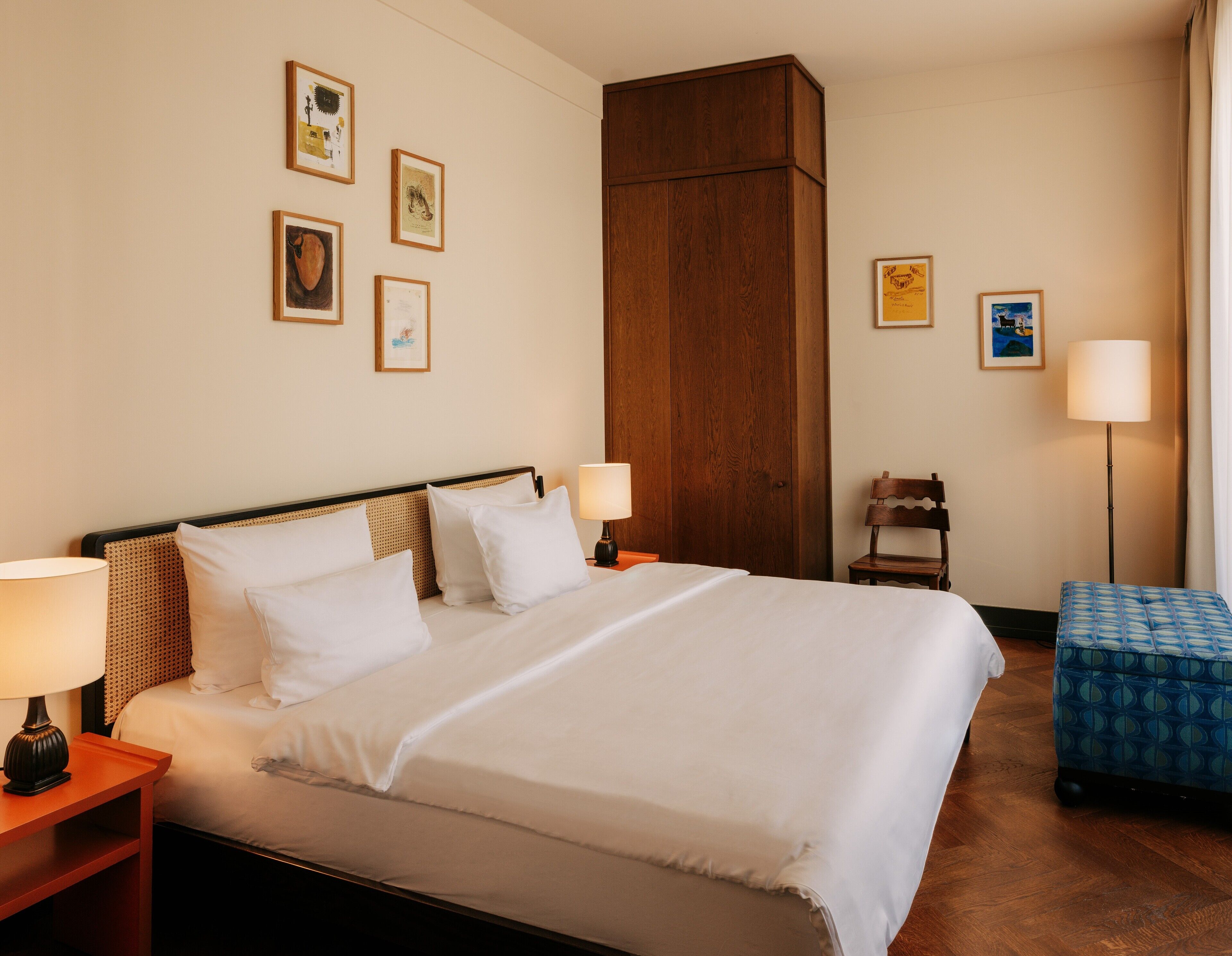 A hotel room with a large bed, two bedside tables, a wooden wardrobe, a small chair, a blue ottoman, and several framed pictures on the wall.