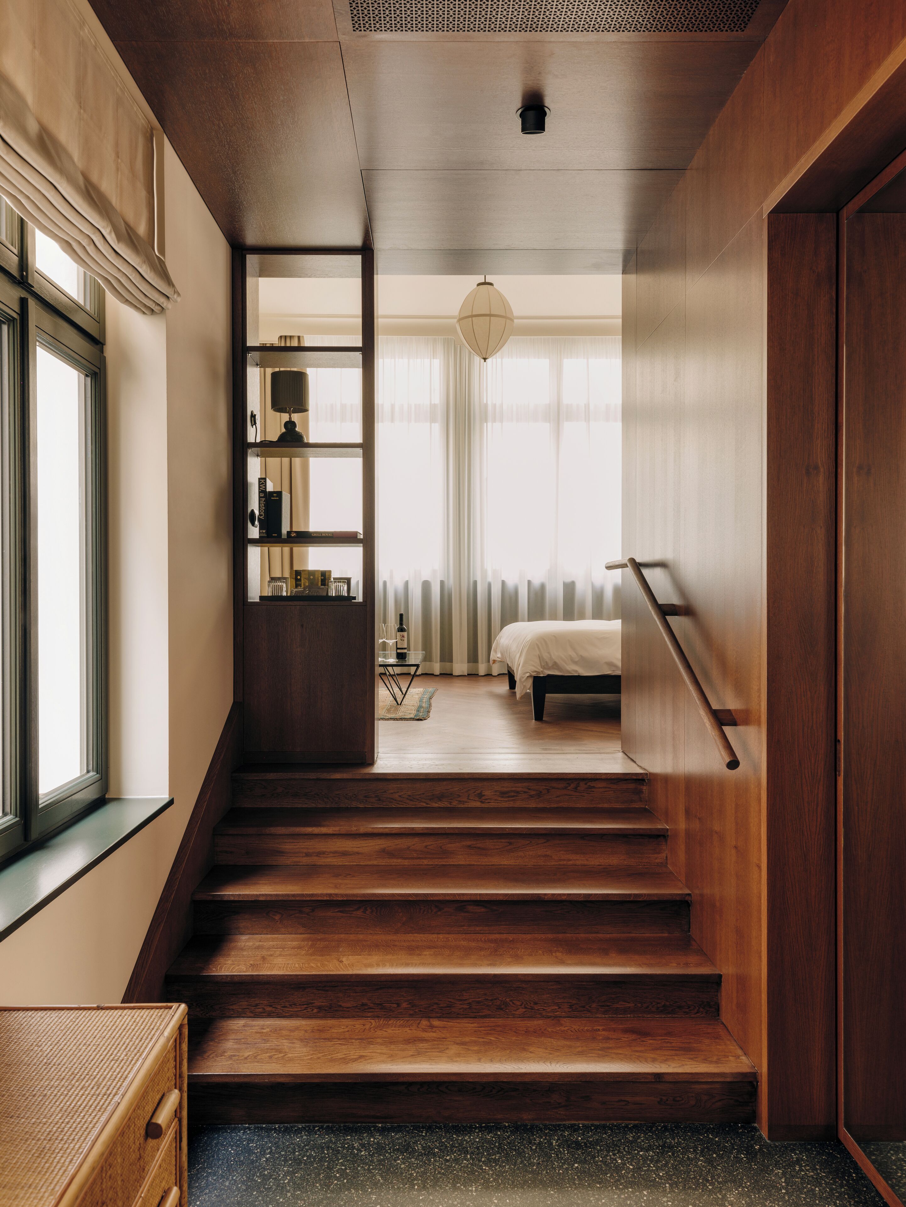 Una escalera moderna conduce a un dormitorio con cama, mesita de noche y lámpara. Se ve una ventana a la izquierda y un radiador en la habitación.