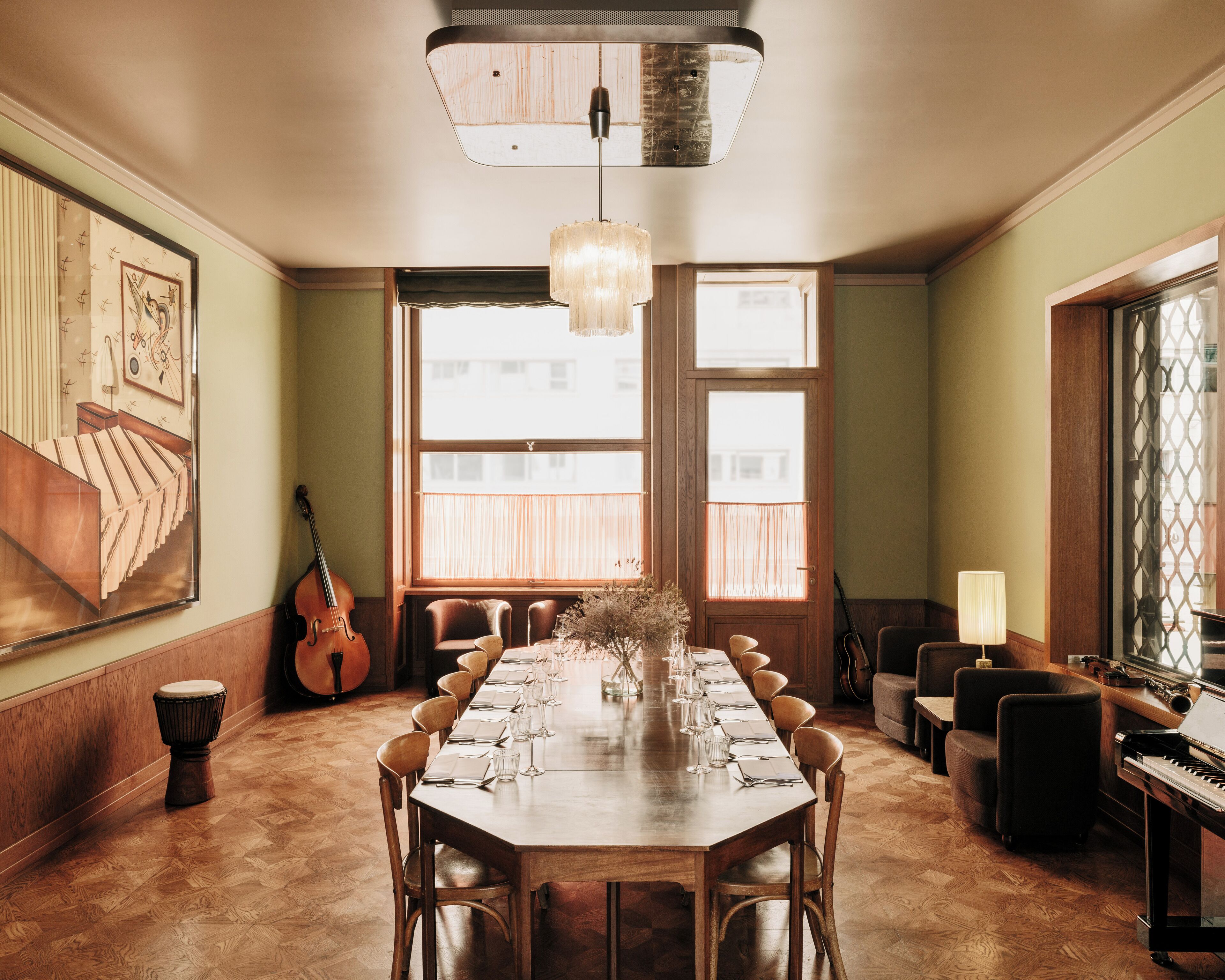 A dining room with a long table set for six, flanked by chairs. A violin is leaning against the wall, and a piano is in the corner.