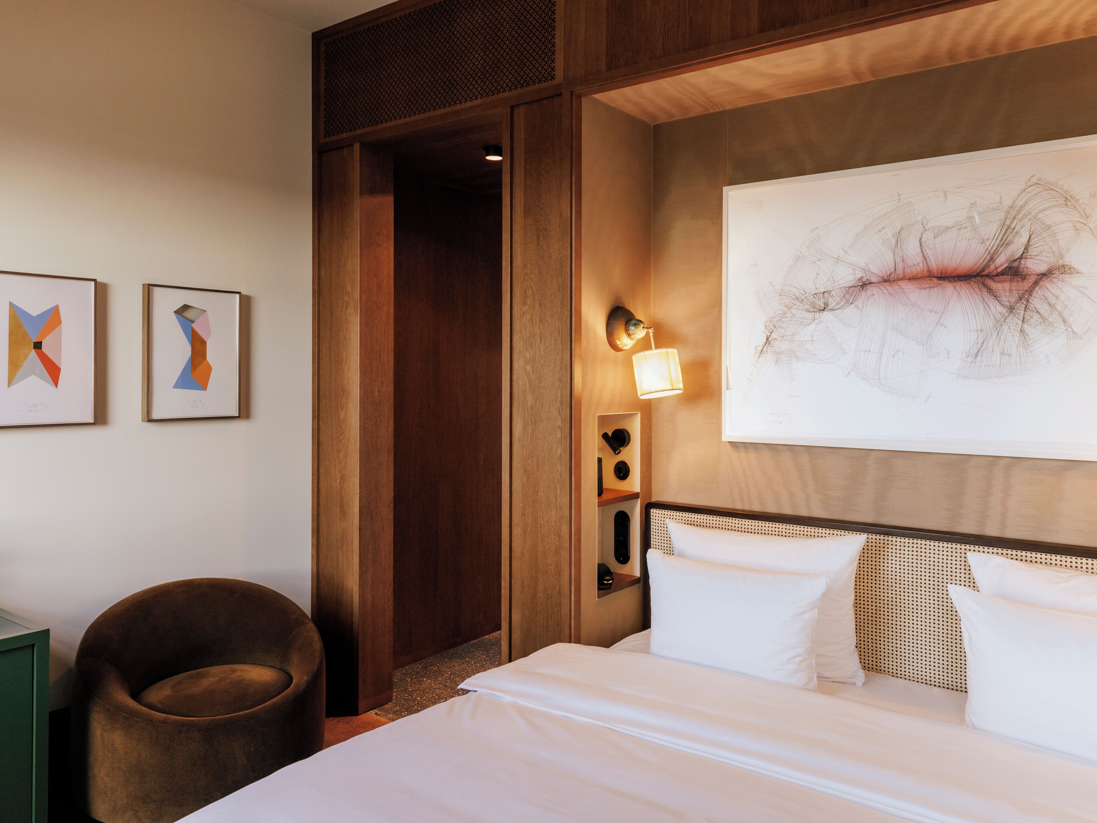 Habitación de hotel con cama, sillón, una obra de arte en la pared y puerta que da a otra habitación.