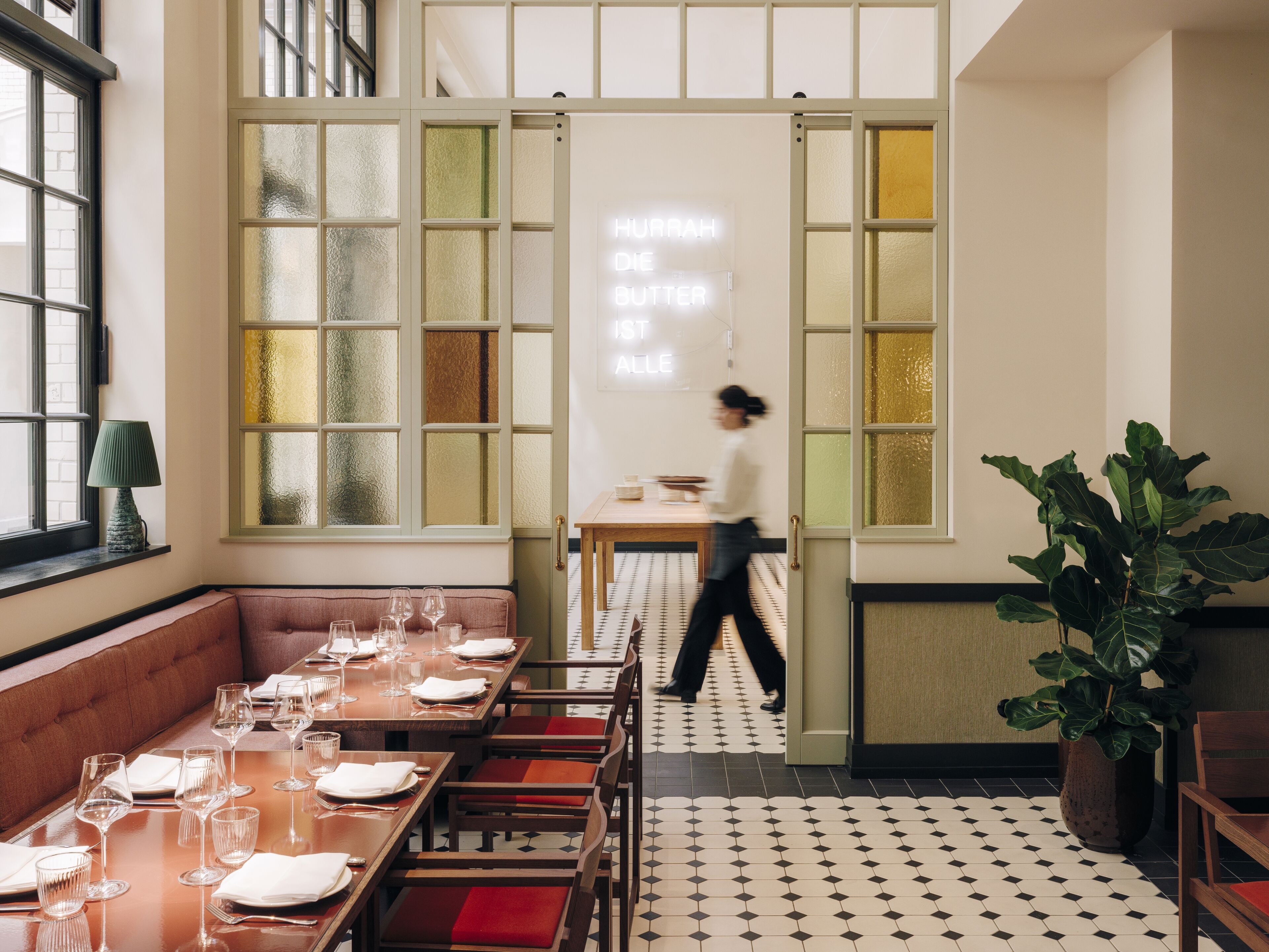 Un restaurante con una decoración moderna, que cuenta con una zona de comedor con mesas preparadas para una comida, una planta grande y una persona caminando por el espacio.