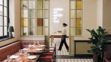 Un restaurante con una decoración moderna, que cuenta con una zona de comedor con mesas preparadas para una comida, una planta grande y una persona caminando por el espacio.