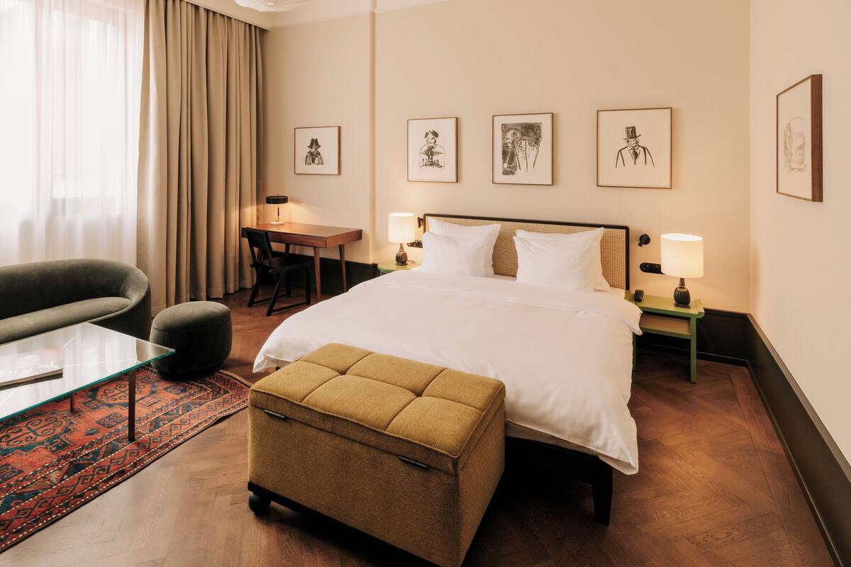 Une chambre d’hôtel avec un lit, un bureau, une chaise, un canapé et un petit pouf. Quatre tableaux encadrés sont accrochés au mur.