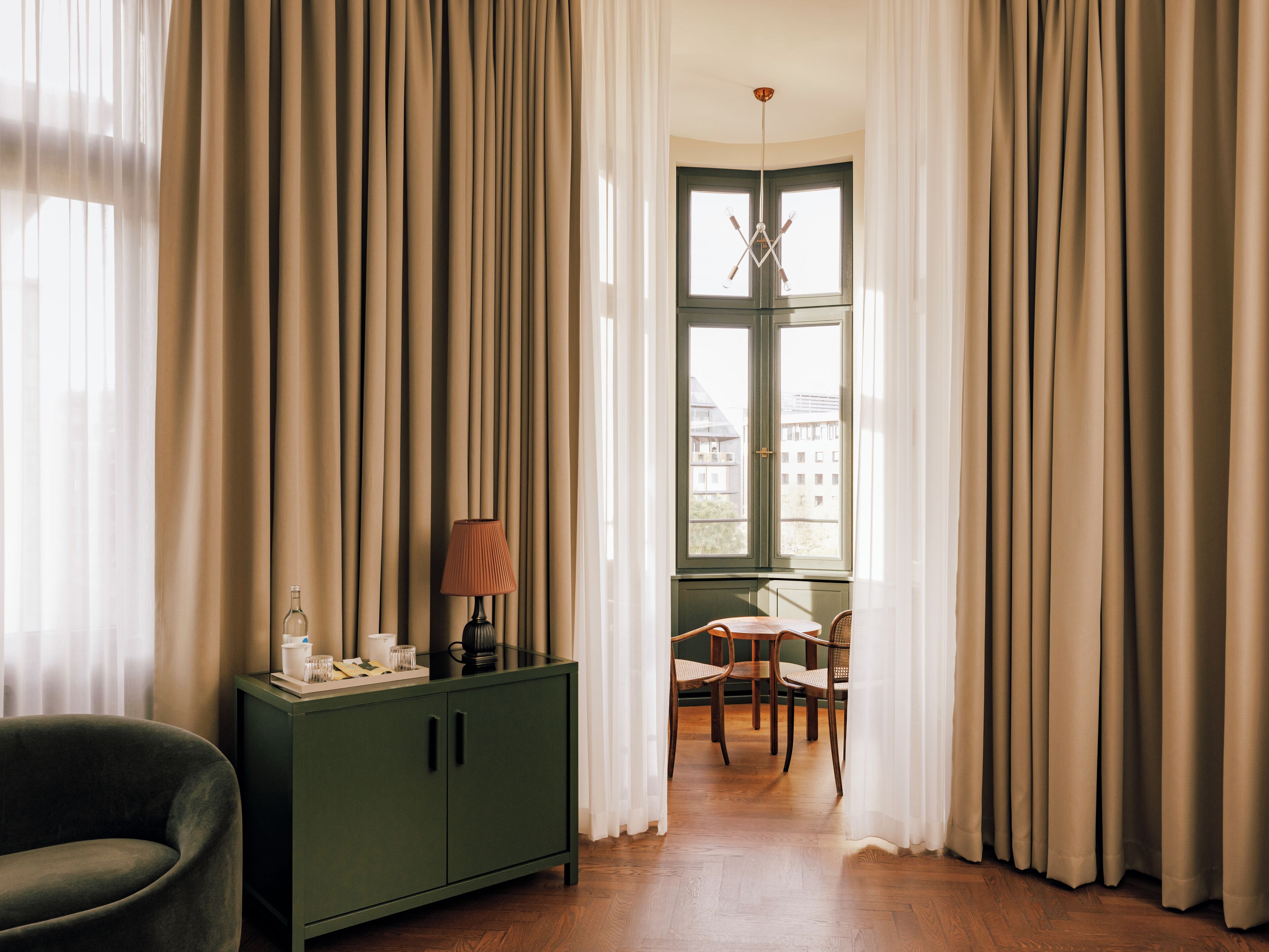 Una habitación con un armario verde, una silla y una mesa con una lámpara. Hay una ventana con cortinas y vistas a edificios.