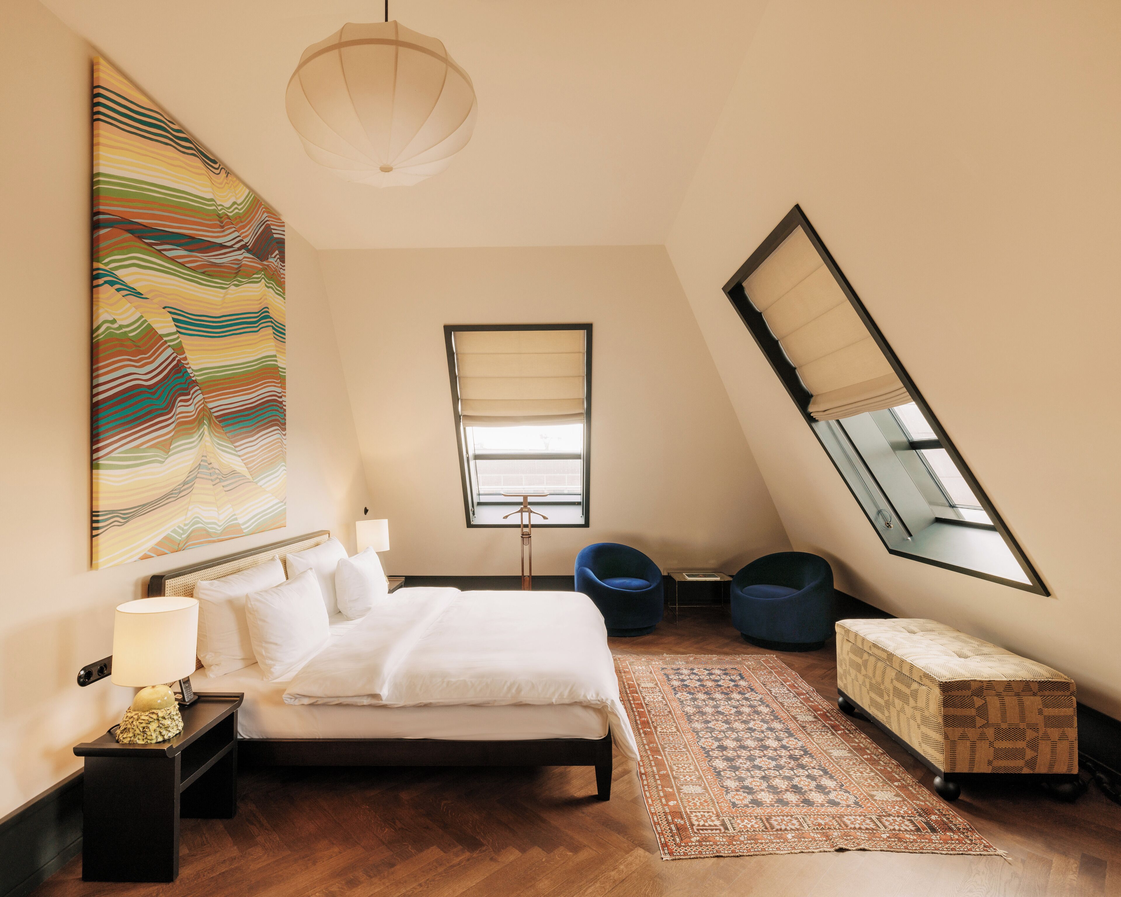 A bedroom with a bed, bedside table, lamp, and a large abstract painting. There is a window with a view of the outside, a hanging light fixture, and a small seating area with a bench and chairs.