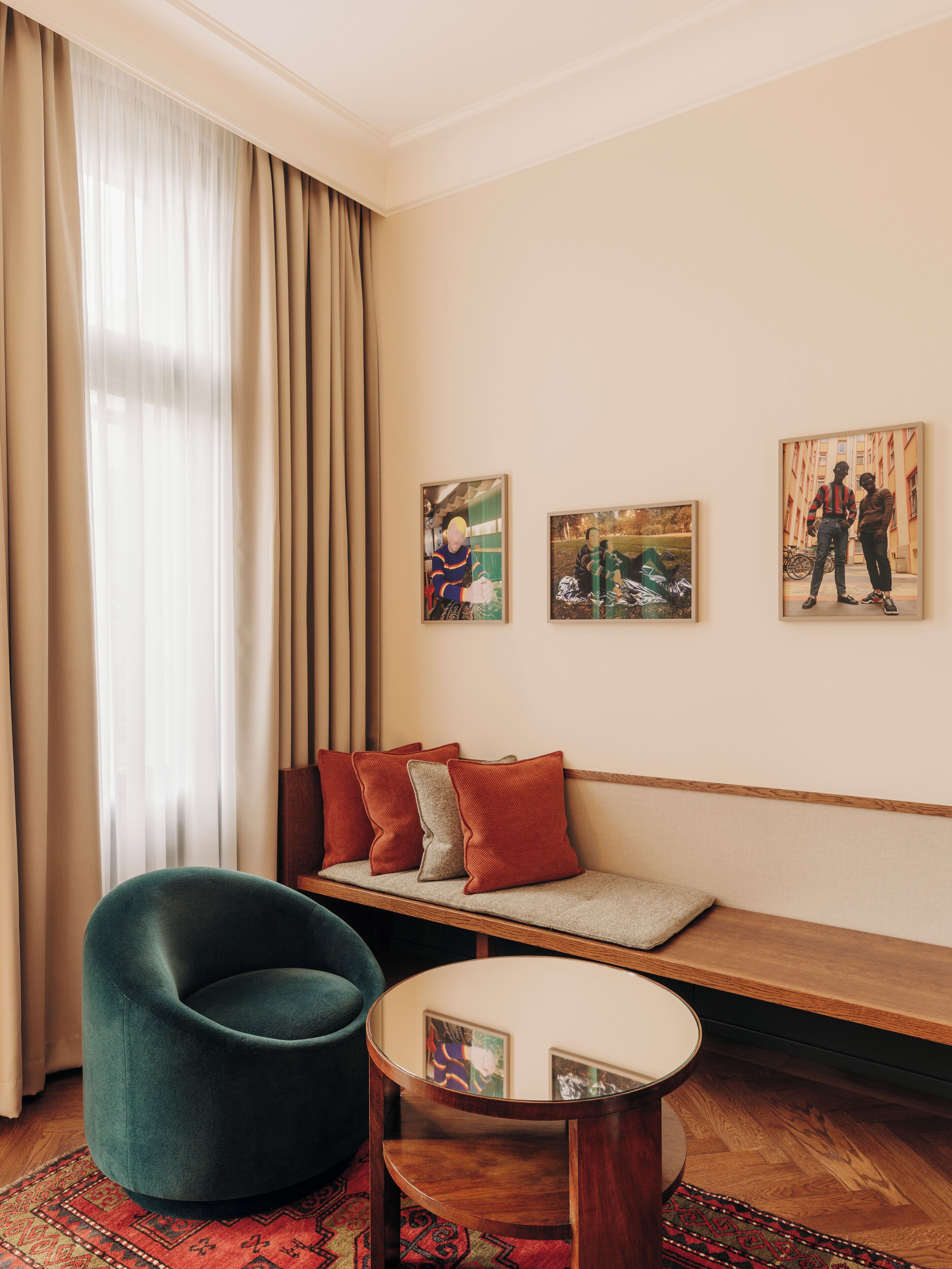 A modern living room with a green armchair, a wooden bench with cushions, and a round wooden table. There are three framed artworks on the wall.