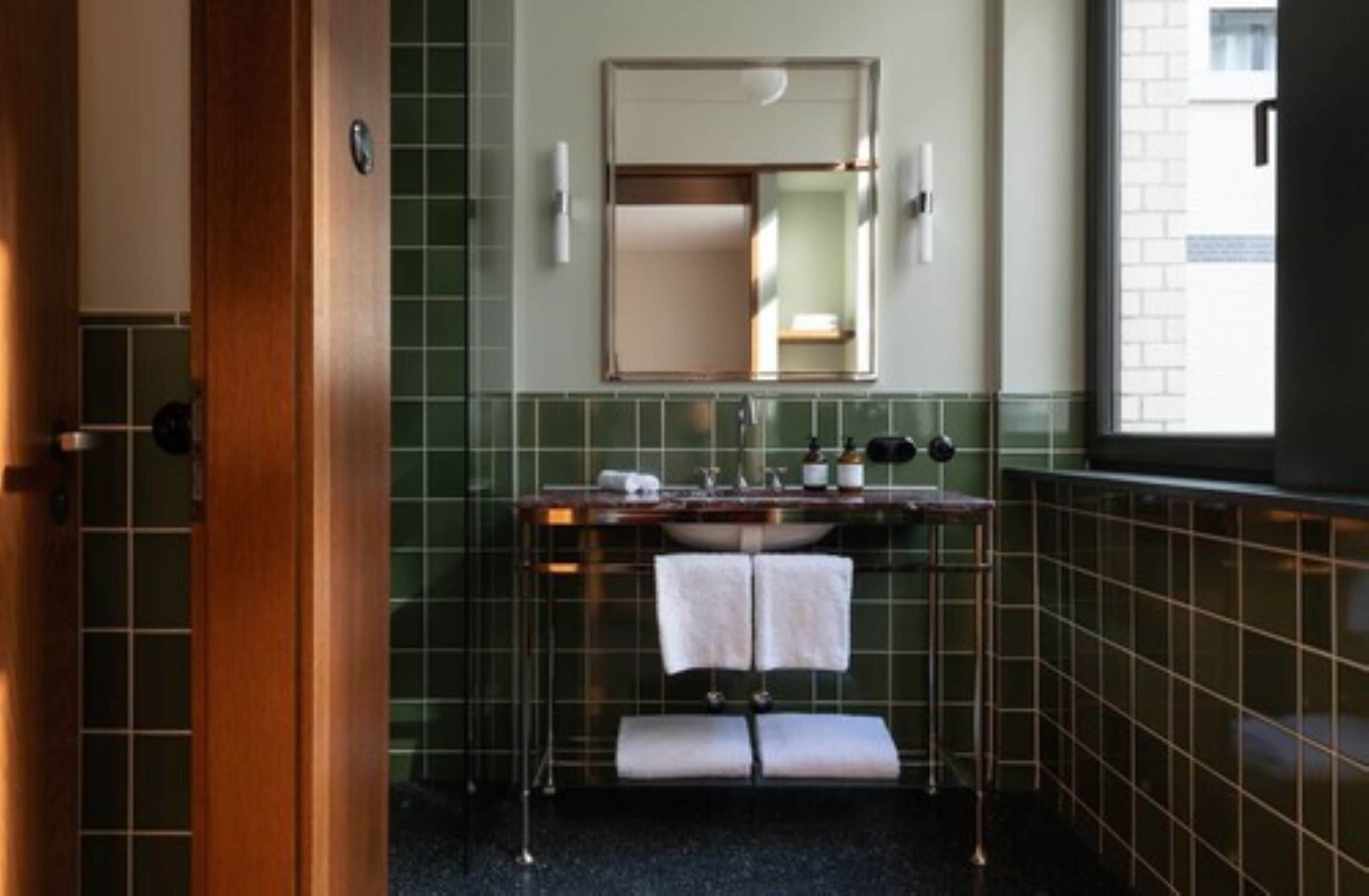 Un baño con puerta de madera, paredes con azulejos verdes, un espejo, un lavamanos con dos toallas y una ventana.