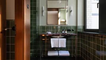 Un baño con puerta de madera, paredes con azulejos verdes, un espejo, un lavamanos con dos toallas y una ventana.