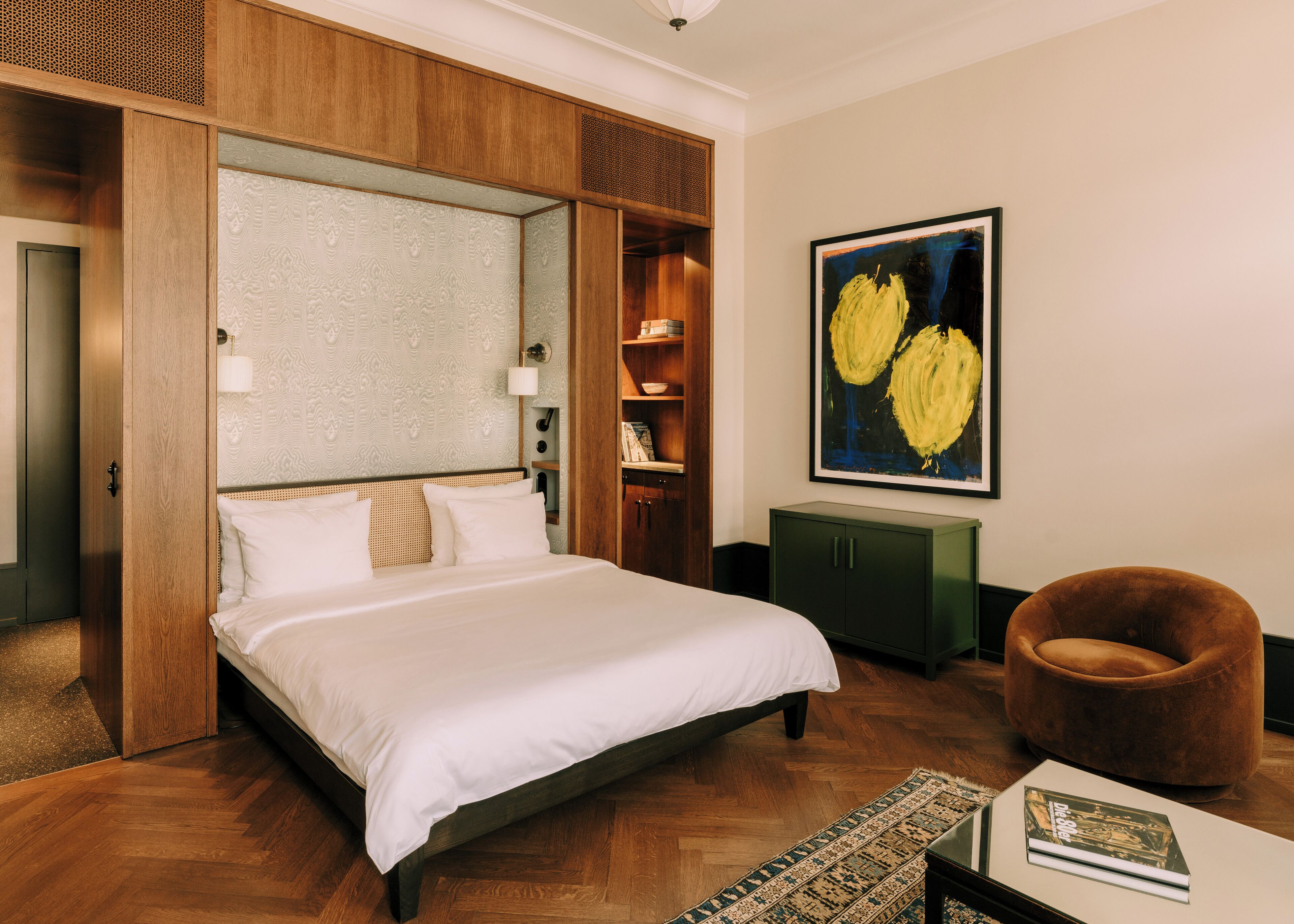 Une chambre d’hôtel avec un grand lit, une armoire en bois, un meuble vert, un fauteuil marron et un tableau au mur.