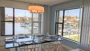 A dining area with a glass table and chairs, large windows with a marina view, and a hanging light fixture. - NEW NEW   Burnt Store Marina Front,  Premier Corner Unit, 30-Day Minimum Retreat (Punta Gorda)