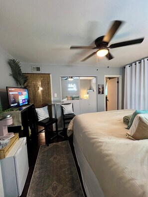Habitación de hotel con cama, escritorio, silla, televisor, ventilador de techo y espejo.
