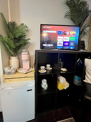 Un salón con televisor en un mueble bajo, un refrigerador pequeño, una cafetera, una planta y un estante con adornos.