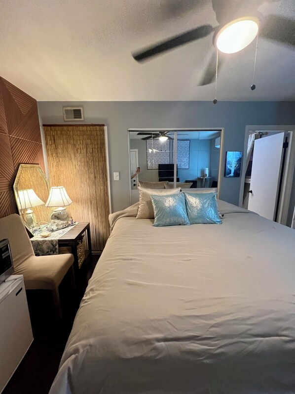 A hotel room with a bed, bedside lamps, a desk with a lamp, a mirror, a ceiling fan, and a view into the adjacent room.