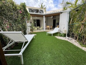 Een achtertuin met kunstgras, twee witte lounge fauteuils en een overdekt terras met een tafel en stoelen.