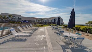 Eine Holzterrasse mit Liegestühlen und einem Tisch mit Stühlen, mit Blick auf einen Pool und einen Strandbereich mit Gebäuden im Hintergrund.